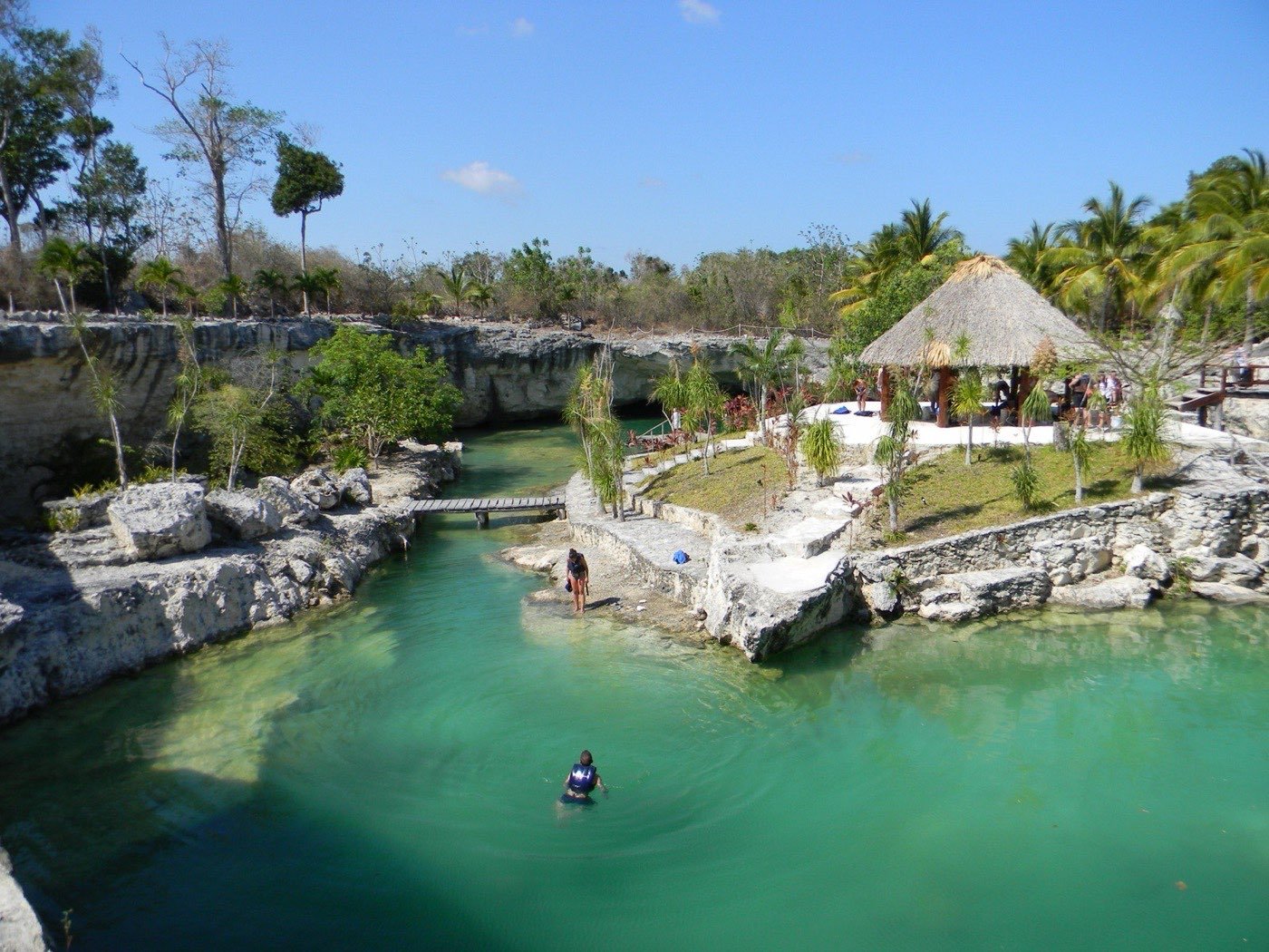 Cenote abierto con palapa y bañistas relajados en aguas turquesas poco profundas — ideal para familias
